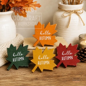 May include: Four wooden maple leaf cutouts in green, yellow, orange, and red, each featuring the white printed text hello AUTUMN. The leaves are arranged on a wooden surface with a white vase and a pinecone visible in the background.