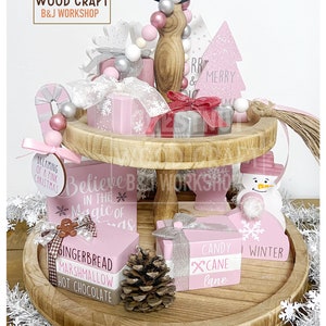 May include: A two-tiered wooden tray with Christmas-themed decorations. The tray holds pink and silver gift boxes, a pink Christmas tree, and a snowman. Text includes "Believe in the Magic of Christmas" and "Candy Cane Lane."