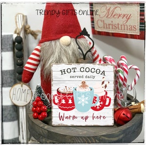 May include: A festive holiday display featuring a gnome with a red hat and striped arms, a wooden sign that reads "Hot Cocoa served daily, Warm up here", and mugs of hot cocoa. Candy canes and a red bell add to the Christmas theme.