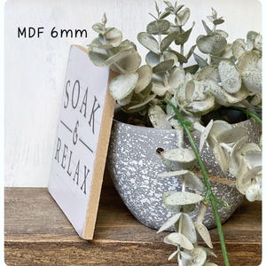 May include: A white MDF sign with the words "SOAK & RELAX" in black lettering. The sign is 6mm thick and is next to a gray speckled pot filled with faux eucalyptus. The sign is on a wooden surface.
