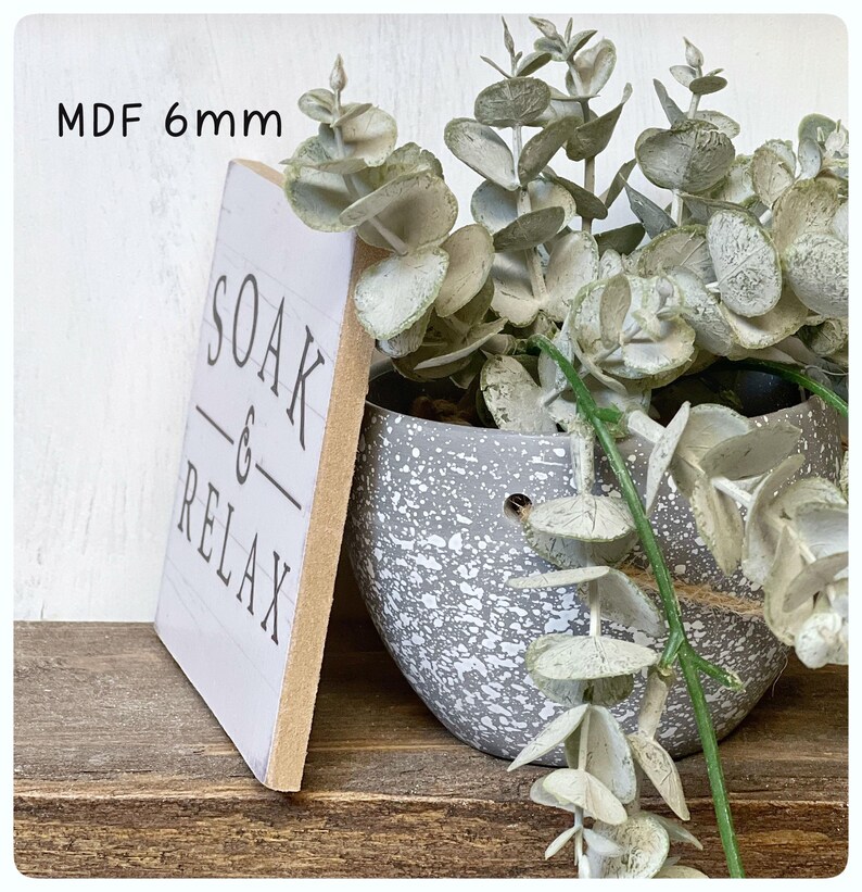 May include: A white MDF sign with the words "SOAK & RELAX" in black lettering. The sign is 6mm thick and is next to a gray speckled pot filled with faux eucalyptus. The sign is on a wooden surface.