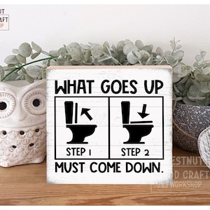 May include: A white wooden sign with black text that reads "What goes up must come down." The sign has two illustrations of toilets, one with an arrow pointing up and the other with an arrow pointing down. The text "Step 1" is below the first illustration and "Step 2" is below the second illustration.