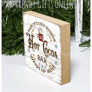 May include: A rectangular wooden sign with a distressed white front and brown sides. The sign reads "Baby it's cold outside, Hot Cocoa Bar, Warm up here" in brown and gold lettering, with a red cocoa cup graphic.