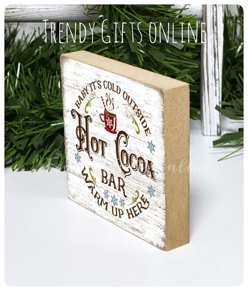 May include: A rectangular wooden sign with a distressed white front and brown sides. The sign reads "Baby it's cold outside, Hot Cocoa Bar, Warm up here" in brown and gold lettering, with a red cocoa cup graphic.