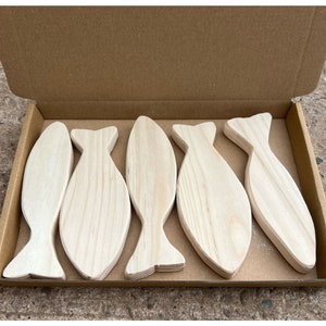 May include: Five unfinished light-coloured wooden fish cutouts arranged side by side inside a rectangular brown cardboard box. Each wooden fish shape features a smooth surface with visible natural wood grain patterns and a simple tail design.