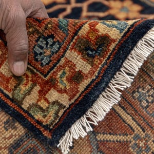 May include: Close-up of a section of a patterned rug. The rug features a complex design with a navy blue border and a cream-colored fringe. The pattern includes shades of red, brown, and blue, creating a detailed and textured appearance.