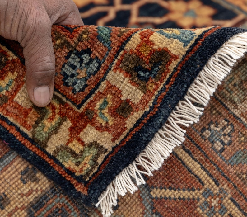 May include: Close-up of a section of a patterned rug. The rug features a complex design with a navy blue border and a cream-colored fringe. The pattern includes shades of red, brown, and blue, creating a detailed and textured appearance.