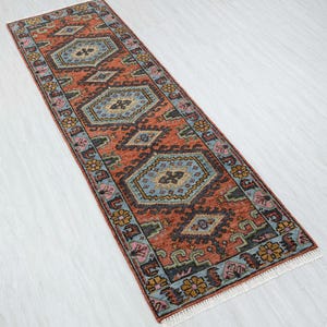 May include: A rectangular runner rug with an intricate geometric pattern. The rug features a burnt orange background with repeating blue, black, and gold diamond and floral designs. The edges are bordered with a floral pattern and a white fringe.