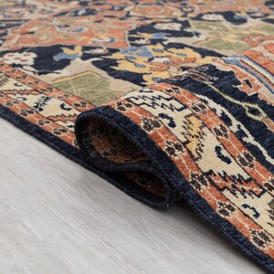 May include: Close-up of a rolled-up area rug with a detailed, traditional design. The rug has a navy blue border and a central pattern in shades of orange, beige, and blue. The rug's intricate pattern is visible, showcasing its craftsmanship.