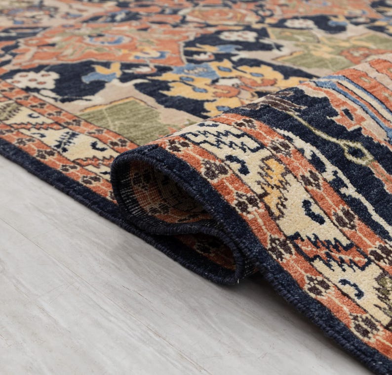 May include: Close-up of a rolled-up area rug with a detailed, traditional design. The rug has a navy blue border and a central pattern in shades of orange, beige, and blue. The rug's intricate pattern is visible, showcasing its craftsmanship.
