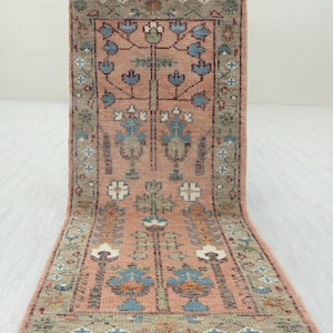 Könnte beinhalten: Ein rechteckiger, handgeknüpfter Teppich mit einem zentralen Blumendesign in Pfirsich-, Blau- und Grüntönen. Der Teppich hat einen detaillierten Rand und Fransen an den Kanten. Die Abmessungen des Teppichs sind auf dem Bild nicht sichtbar.