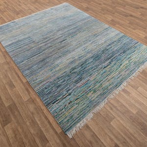 May include: A rectangular area rug with a striped pattern in shades of blue, green, and beige. The rug has a fringe on one end and is made of woven material. The rug is placed on a wooden floor.