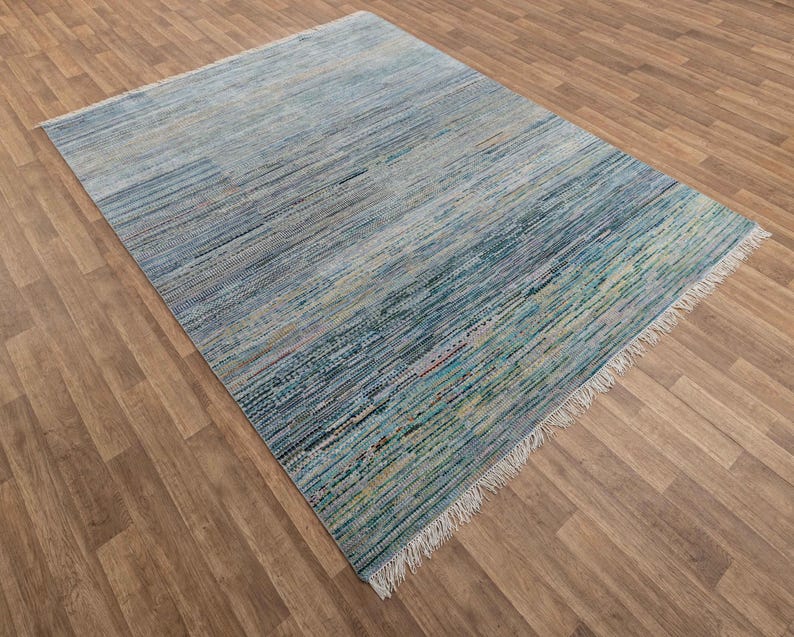 May include: A rectangular area rug with a striped pattern in shades of blue, green, and beige. The rug has a fringe on one end and is made of woven material. The rug is placed on a wooden floor.