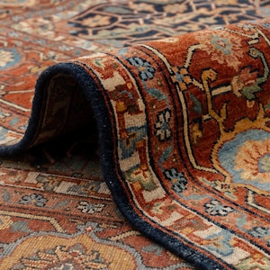 May include: Close-up of a rolled-up, intricately patterned rug. The rug features a rich color palette of rust, navy blue, and beige, with floral and geometric designs. The border is a deep navy blue, and the rug appears to be made of a woven material.