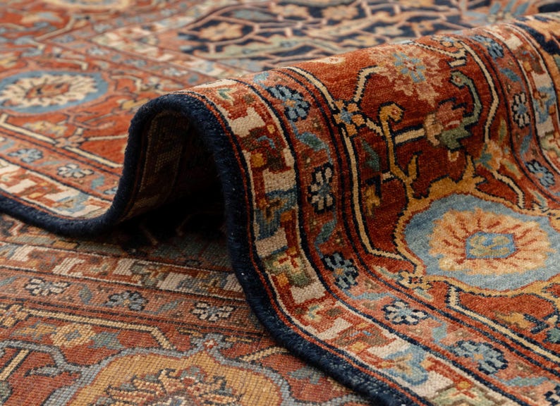 May include: Close-up of a rolled-up, intricately patterned rug. The rug features a rich color palette of rust, navy blue, and beige, with floral and geometric designs. The border is a deep navy blue, and the rug appears to be made of a woven material.