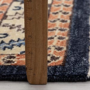 May include: Close-up of a small, rectangular rug with a traditional design. The rug features a cream-colored field with blue and orange geometric patterns. A wooden ruler is placed vertically along the edge, showing measurements.
