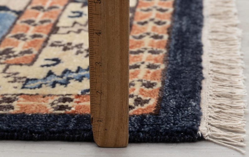 May include: Close-up of a small, rectangular rug with a traditional design. The rug features a cream-colored field with blue and orange geometric patterns. A wooden ruler is placed vertically along the edge, showing measurements.