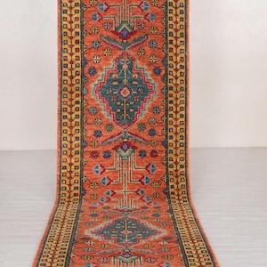 Puede incluir: Una alfombra de pasillo naranja con un patrón geométrico detallado. La alfombra presenta un diseño central en verde azulado, rojo, azul y amarillo, con un borde de formas repetidas. La alfombra es rectangular y parece estar hecha de material tejido.