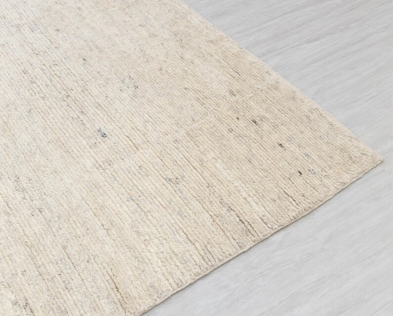 May include: A close-up of a cream-colored rug with a textured, woven surface. The rug has a subtle, natural pattern and a slightly rough texture.