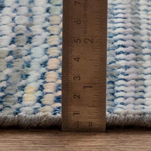 May include: Close-up of a textured rug with a blue, white, and gray pattern. A wooden ruler is placed vertically along the edge of the rug, showing measurements in inches. The rug has a soft, plush appearance.