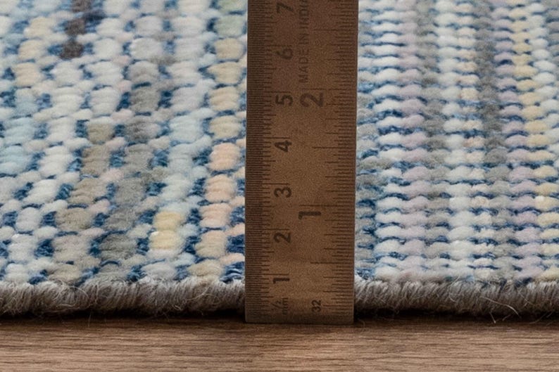 May include: Close-up of a textured rug with a blue, white, and gray pattern. A wooden ruler is placed vertically along the edge of the rug, showing measurements in inches. The rug has a soft, plush appearance.