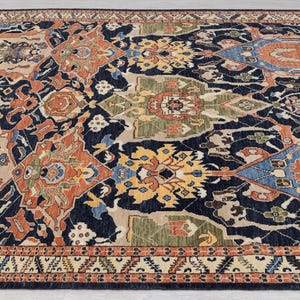 May include: A large, rectangular rug with a detailed, traditional design. The rug has a dark navy blue background with a complex pattern of orange, beige, yellow, and blue geometric shapes. The border has a repeating pattern in beige and orange.