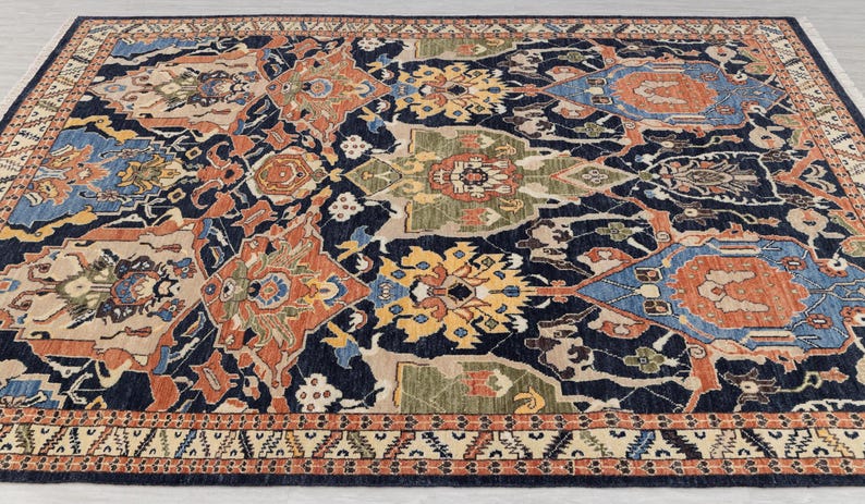 May include: A large, rectangular rug with a detailed, traditional design. The rug has a dark navy blue background with a complex pattern of orange, beige, yellow, and blue geometric shapes. The border has a repeating pattern in beige and orange.