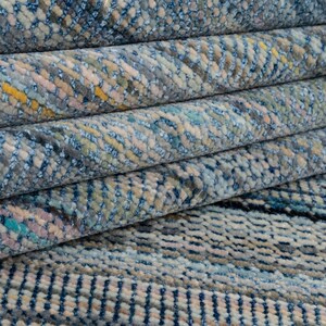 May include: Close-up of a rolled-up rug with a textured pattern. The rug features a mix of blue, gray, yellow, and pink hues. The design appears to be a woven or knotted style, with visible texture and detail. The rug is folded over itself.