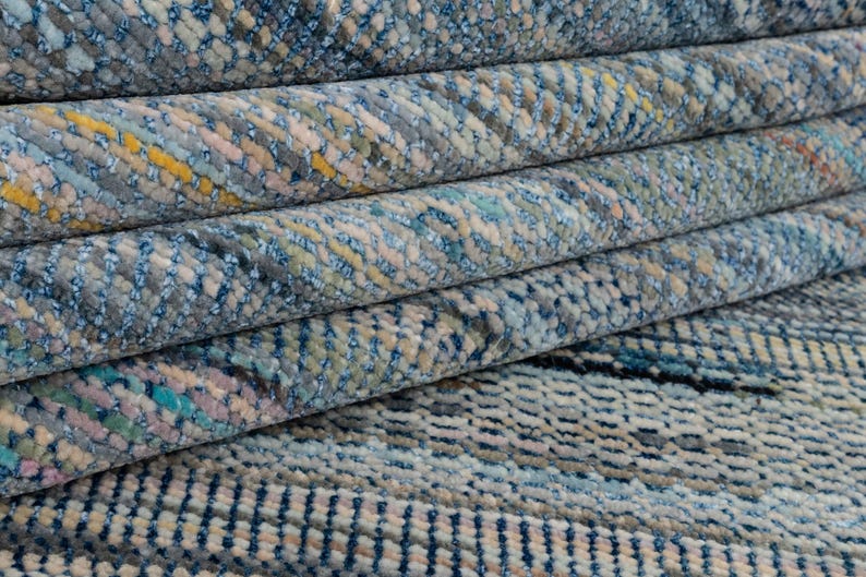 May include: Close-up of a rolled-up rug with a textured pattern. The rug features a mix of blue, gray, yellow, and pink hues. The design appears to be a woven or knotted style, with visible texture and detail. The rug is folded over itself.