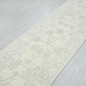 May include: Cream-colored runner rug with a floral pattern in light blue and beige. The rug has a rectangular shape and is placed on a light gray surface. The design features a central floral motif and a border.