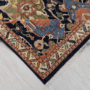 May include: Detail of a patterned rug with a navy blue border and a cream and orange geometric design. The central pattern features blue, orange, green, and beige floral motifs. The rug is folded.