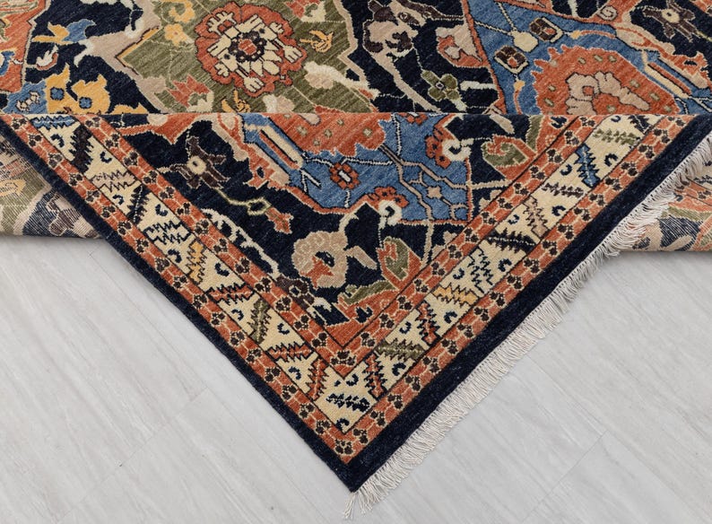 May include: Detail of a patterned rug with a navy blue border and a cream and orange geometric design. The central pattern features blue, orange, green, and beige floral motifs. The rug is folded.