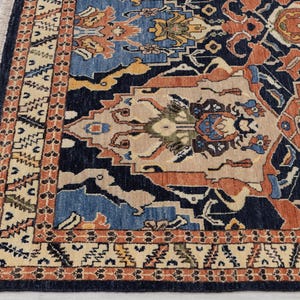 May include: Detail of a patterned rug with a navy blue base and intricate designs in blue, orange, beige, and yellow. The border features a repeating geometric pattern. The rug has a fringed edge. The rug's design is complex.