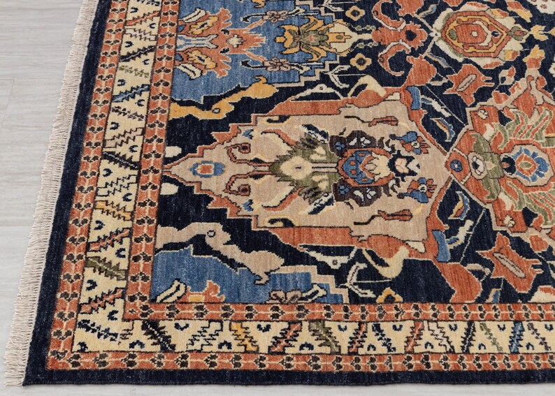 May include: Detail of a patterned rug with a navy blue base and intricate designs in blue, orange, beige, and yellow. The border features a repeating geometric pattern. The rug has a fringed edge. The rug's design is complex.