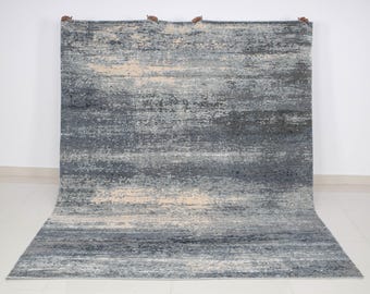 Puede incluir: Una alfombra rectangular grande con un diseño abstracto texturizado. La alfombra presenta una mezcla de tonos grises, azules y beige, creando un efecto acuarela en capas. La alfombra se muestra contra una pared blanca, mostrando sus colores y diseño.