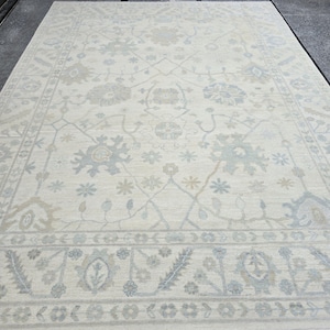 May include: A light beige rug with a delicate floral pattern in shades of blue and gray. The rug has a border with a repeating pattern of small flowers and leaves.