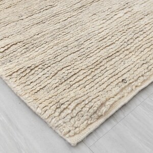 May include: Close-up of a beige, textured rug with a slightly frayed edge. The rug has a woven, hand-crafted look.
