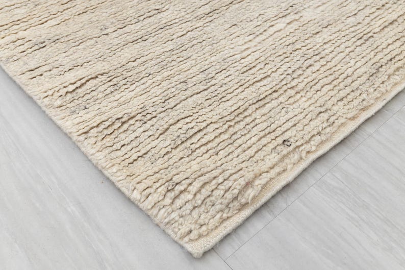 May include: Close-up of a beige, textured rug with a slightly frayed edge. The rug has a woven, hand-crafted look.