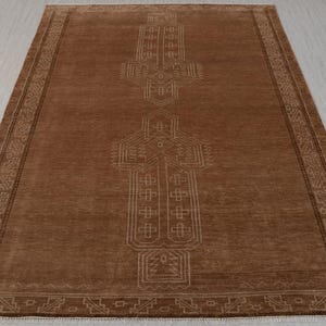 May include: A brown area rug with a geometric pattern in a lighter brown color. The pattern is centered and features a repeating design of squares and diamonds.