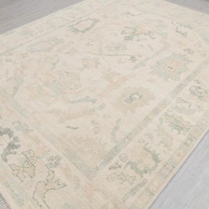 Op de afbeelding: Een rechthoekig, gebroken wit tapijt met een gedetailleerd bloemenpatroon in beige en lichtgroen. Het tapijt heeft een rand met een vergelijkbaar ontwerp en een gefranjerde rand. Het ligt op een lichtgrijze ondergrond.