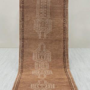 Puede incluir: Una alfombra de pasillo rectangular marrón con un patrón geométrico detallado en un tono más claro de marrón. La alfombra tiene flecos en ambos extremos y mide aproximadamente 91 cm de ancho.