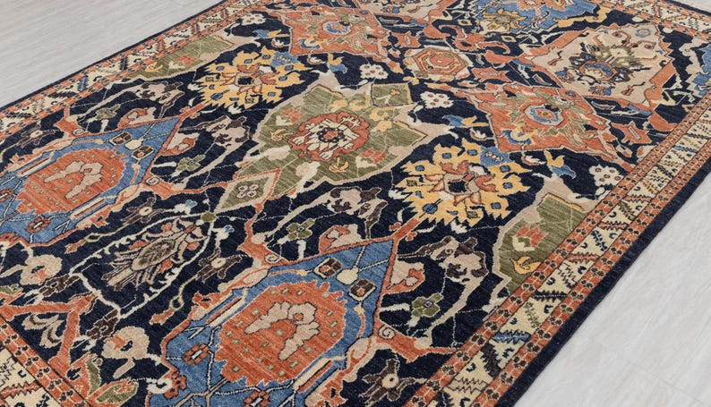 May include: A rectangular rug with a detailed, traditional design. The rug has a dark navy blue background with a repeating pattern of floral and geometric shapes in shades of orange, blue, green, and yellow. The border has a cream and brown geometric pattern.