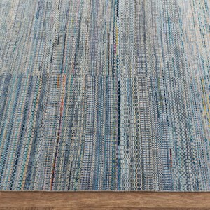 May include: A close-up of a woven rug with a blue and white color scheme. The rug has a textured appearance with vertical stripes and a mix of colors, including blue, white, and hints of orange and yellow. The rug is made of a durable material.