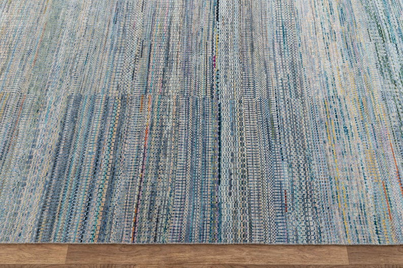 May include: A close-up of a woven rug with a blue and white color scheme. The rug has a textured appearance with vertical stripes and a mix of colors, including blue, white, and hints of orange and yellow. The rug is made of a durable material.