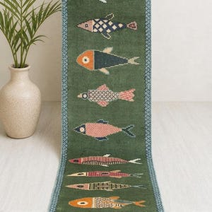 May include: A long, rectangular green runner rug featuring a repeating pattern of stylized fish in various colors including orange, pink, and blue. The rug has a decorative blue border with a geometric pattern and fringed ends at the top and bottom.