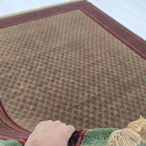 May include: A rectangular area rug featuring a repeating diamond pattern in muted green and reddish-brown tones. The rug is framed by a wide, dark red border with intricate geometric detailing and finished with light beige fringe at the edge.