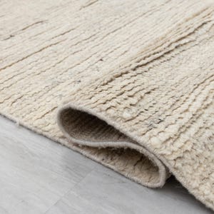May include: Close-up of a cream-colored rug with a textured, woven design. The rug is folded over, showing the edge and the underside of the fabric.