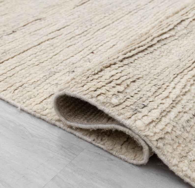 May include: Close-up of a cream-colored rug with a textured, woven design. The rug is folded over, showing the edge and the underside of the fabric.