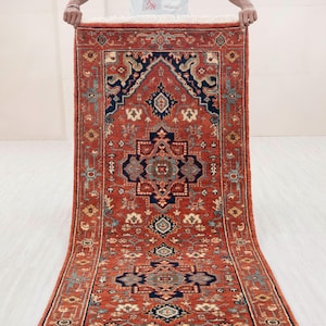 Puede incluir: Una alfombra oriental rectangular roja con un patrón simétrico detallado. La alfombra presenta un diseño de medallón central en azul marino, verde azulado y beige, rodeado de intrincados motivos florales y geométricos. El borde de la alfombra es de un rojo más oscuro.