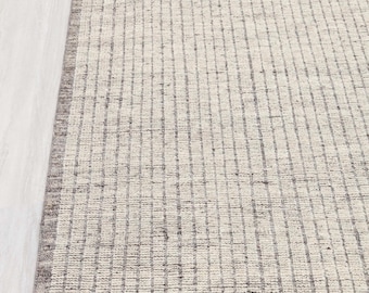 Pode incluir: Um tapete retangular com um padrão listrado texturizado em tons neutros. O tapete apresenta um campo bege claro com listras cinza mais escuras e uma borda correspondente. O design é simples e moderno, adequado para vários estilos de interiores.